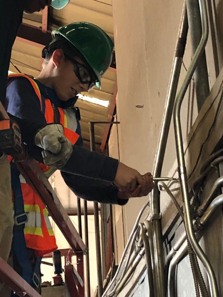 A T.A.C.T. student in protective glasses and hard hat holds a tool.