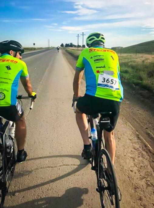 Two cyclists with Sturgeon Electric jerseys riding along the side of a road