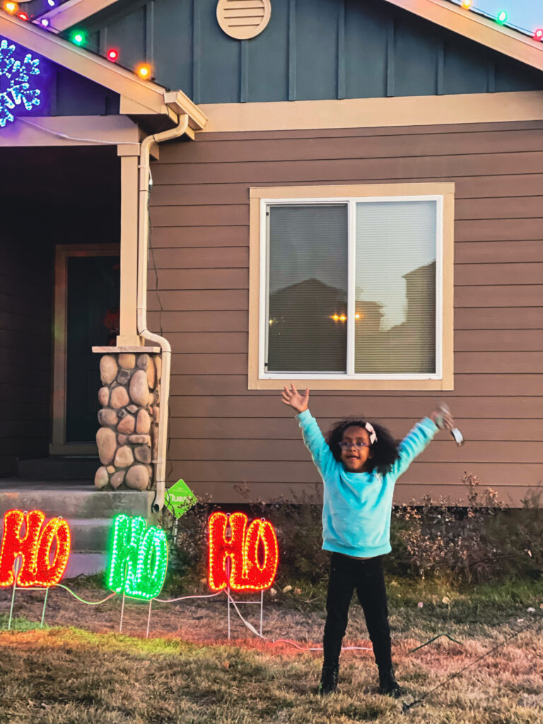 A little girl stands outside in front of festive lights with her arms raised